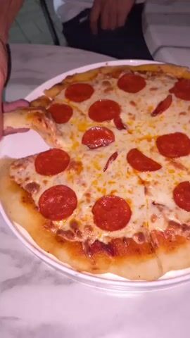 A close-up shot focuses on a pepperoni pizza served on a white plate. The pizza has a golden-brown crust and is topped with melted cheese and numerous slices of pepperoni. A hand is seen reaching into the frame and lifting a slice of pizza, revealing a string of melted cheese connecting it to the rest of the pizza. The background is slightly out of focus, showing parts of what appears to be a dining setting with a marble-patterned tabletop and the blurred figures of people. The overall lighting suggests an indoor environment.