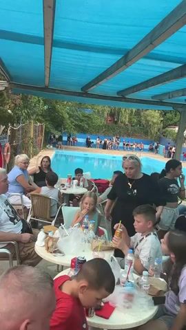 The video shows an outdoor gathering of people, likely at a resort or water park, with a swimming pool visible in the background. The foreground is dominated by tables with people sitting and eating. A woman in a black t-shirt with blonde hair is standing and talking to a young boy. Other people, including children and adults, are seated at tables, some eating and drinking, others looking around. The area is covered by a blue canopy supported by wooden beams. The atmosphere appears casual and relaxed.
