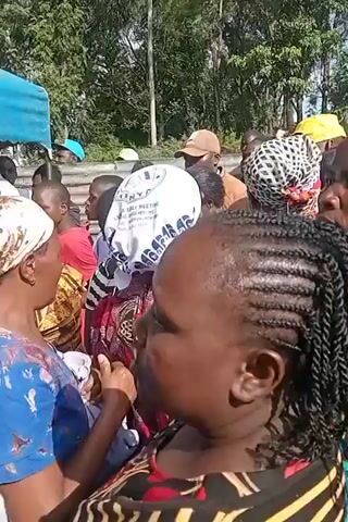 The video shows a crowd of people gathered outdoors, some under a blue and white tent. The lighting suggests it is daytime. The people appear to be of African descent and are dressed in casual clothing. Their faces are mostly turned away from the camera, making it difficult to ascertain their expressions or actions. There are trees and foliage visible in the background, indicating a natural environment. The camera movement is shaky, suggesting it is handheld and possibly moving through the crowd. There is no discernible audio content in the provided frames.