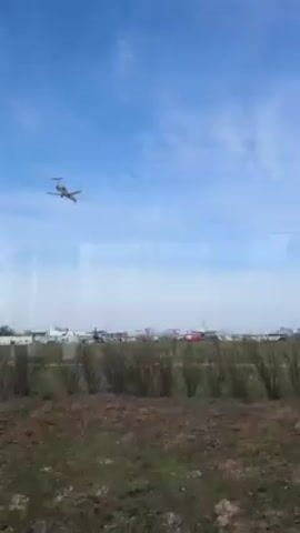 The video, apparently shot through a window, shows an airplane in the process of landing on a runway. The plane is airborne, but clearly descending towards the ground.  Below it is a patch of green grass, then a row of short bushes, followed by brown dirt or soil in the foreground.  In the background, the edge of an airport can be seen, including several other parked planes and some buildings. The sky is blue with light clouds. The reflection of what seems to be the interior of the space where the camera is located is visible on the window.