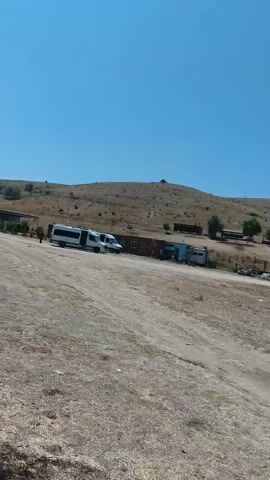 A wide-angle shot shows a rural landscape under a clear blue sky. In the foreground, dry, dusty ground slopes upwards. In the midground, a cluster of small buildings and structures are visible. To the left, a white bus and two white vans are parked on a dirt road. Adjacent to them are several portable buildings or shipping containers, one red, one light blue, and others with darker exteriors. Further to the right, there is a small, stone building with a red roof. Trees are scattered throughout the midground and background. The background consists of rolling, dry, grassy hills dotted with sparse vegetation. The camera pans slowly to the right, revealing more of the landscape and structures.