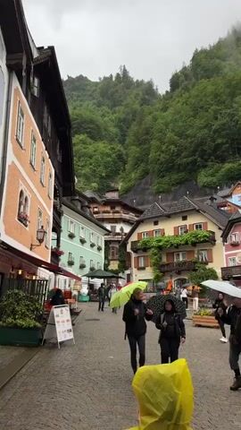 The video captures a town square in Hallstatt, Austria, on a rainy day. The scene is dominated by colorful, traditional Austrian buildings nestled against a lush green mountainside covered in trees. Several people are present, holding umbrellas of various colors, including green, white, and blue, as they walk through the cobblestone square. The weather appears overcast and misty, contributing to the atmospheric feel of the location. Some of the buildings have signs indicating businesses such as "Restaurant" and "Cafe." In the center of the square, there is a stone fountain structure. The overall impression is one of a charming European village experiencing a typical rainy day.