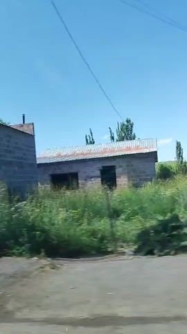 This is a video captured from a moving vehicle, likely a car, as indicated by the motion blur and the perspective. The scene is an outdoor environment with a building visible in the first two frames and trees and vegetation in the subsequent frames.

In the initial frames, a building constructed with greyish-brown stones is prominently featured. It has a large, dark-colored double door with decorative elements and a small window to the left. In the second frame, the building appears to have signage that reads "PLATON DENTW". There's also an air conditioning unit mounted on the exterior wall and another window. The ground in front of the building is paved with light-colored interlocking stones.

As the video progresses, the view shifts to a roadside setting. Large trees with lush green foliage line the path, creating a dappled sunlight effect. A rough stone wall is visible bordering the road, and in one frame, a bright green metal door or gate is partially seen. The sky is a clear blue, suggesting a sunny day. The overall impression is a drive through a semi-urban or rural area with some commercial establishments and natural greenery. The time of day appears to be daytime due to the bright sunlight. No people are clearly visible in the video, and the focus is on the exterior environment and the building.