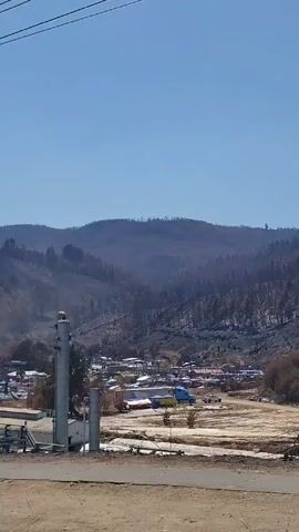 The video appears to be taken from a moving vehicle, possibly a car or train, showing an industrial area against a backdrop of hills or mountains. In the foreground, there is a chain-link fence, a gray building and some industrial equipment, including large white tanks and a tall tower with the name "INDURA" visible on its side.  The background shows what seems to be an area of hillside, potentially scorched by fire. The sky is clear and blue. The camera pans slightly to the right, maintaining focus on the industrial plant as it moves past. Power lines are visible stretching across the top of the frame.
