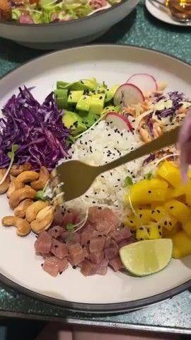 A close-up shot shows a poke bowl being mixed with a fork. The bowl contains rice, diced tuna, avocado, red cabbage, cashews, mango, and lime. A hand with manicured nails is holding the fork and is in the process of combining the ingredients. The background is slightly blurred, showing another plate with food. The lighting is bright, and the colors of the food are vibrant.