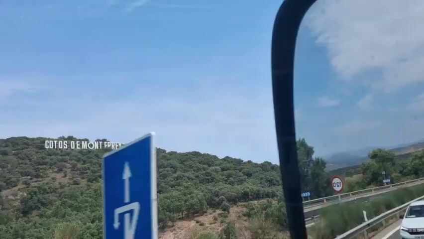 The video shows a landscape with a sign on a hill that reads "COTOS DE MONT ERREY". The hill is covered with trees and shrubs. There is a blue sky with some clouds. A portion of a vehicle interior is visible in the foreground.