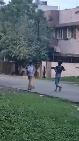 A group of young people are playing cricket on a paved road. Two individuals are positioned near a makeshift wicket. One person stands holding a cricket bat, appearing ready to bat, while another individual is a short distance away, also holding a bat. A third person is visible further down the road, walking away from the scene. The environment appears to be a residential area with buildings and trees in the background. The ground is paved and there is grass on the side of the road. The lighting suggests it is daytime, possibly late afternoon or early evening.