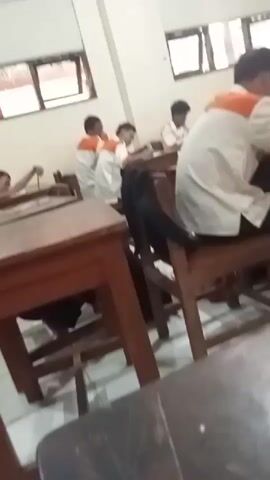 The video shows a classroom setting with several students seated at desks. The students are wearing white shirts with orange accents. One student in the foreground is writing on a piece of paper. Another student to his left is also writing. A student in the background is looking down at his desk. The desks are made of wood and appear to be individual student desks. The walls of the classroom are painted a light color, and there are windows with blinds visible. The lighting in the room is dim.
