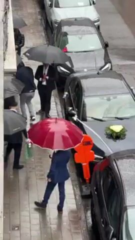 Here is a detailed description of the video:

**Scene:** A wedding ceremony is taking place on a wet day in a narrow street or alley.

**Environment:** The street is paved and visibly wet from the rain. There are buildings lining one side of the street.

**Objects:**
*   Several cars are parked along the street. At least one of the cars, a black sedan, has a small floral arrangement on the hood, indicating it may be a wedding car.
*   People are holding umbrellas. The umbrellas are mostly black, but one distinctive umbrella is red.
*   There is an orange safety cone or construction marker positioned on the street next to one of the cars.

**People:**
*   Several individuals are present, mostly dressed in suits or formal attire. These individuals are presumed to be wedding guests.
*   Many are carrying umbrellas.

**Actions:**
*   People are walking and standing around.
*   The rain is falling heavily.

**Interactions:**
*   People are using umbrellas to shield themselves from the rain.
*   The presence of formal wear and a wedding car suggests that the people present are attending a wedding.
*   A person seems to be carrying a green bag that looks like a gift.

**Summary:** The video depicts a rainy wedding scene on a street. Wedding guests, dressed in formal wear, are using umbrellas to protect themselves from the rain while walking around.