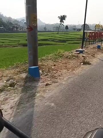 The video shows a view from a motorcycle as it travels along a paved road. To the left of the road are bright green rice paddies, bordered by dirt and some construction materials, including blue blocks and bricks. A large sign with red and white lettering is visible, spelling out "BENDUNGAN GAYAR" in large, block letters. The sign has yellow lettering at the top that is partially obscured. Behind the rice paddies, a lush green landscape with trees and rolling hills can be seen under a bright sky. The motorcycle's handlebar and mirror are visible in the foreground. The camera is moving, suggesting a journey.