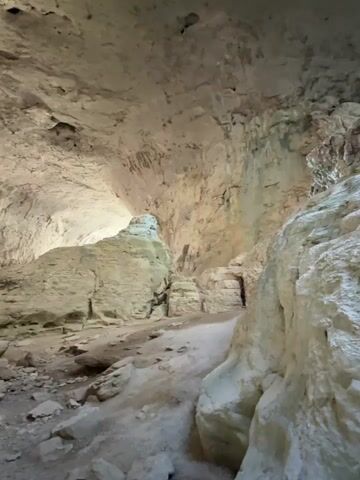 The video pans slowly across the interior of a large cave. The walls and ceiling are rough, textured rock in shades of tan and white, with visible mineral deposits and natural formations. Sunlight, or artificial light, illuminates the cavern from an unseen source, creating shadows and highlighting the contours of the rock. In the foreground, loose rocks and sandy, uneven ground cover the cave floor. A prominent, large rock formation stands in the center of the frame, with smaller rock piles and debris scattered around it. A narrow passageway is visible to the right, leading further into the darkness of the cave. The overall impression is of a natural, ancient geological space.