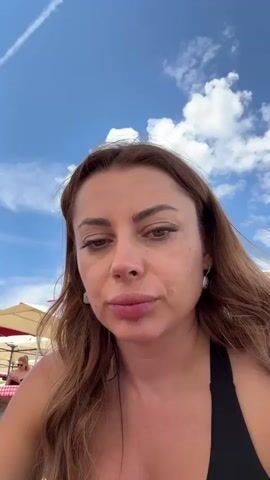 A woman with long, auburn hair and light makeup is sitting outdoors, possibly at a cafe or restaurant, as indicated by the tables and umbrellas in the background. She is wearing a black top and is looking directly at the camera. She brings a forkful of food to her mouth and eats it. The sky is blue with white clouds.