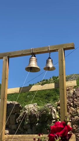A short video captures a serene scene with two brass bells hanging from a wooden structure against a clear blue sky. The bells are being rung, indicated by the faint motion blur of the bell clappers. In the foreground, vibrant red roses in full bloom add a splash of color, with green leaves and stems visible. The background reveals a rocky, hillside landscape covered in lush green vegetation, suggesting a mountainous or hilly terrain. A stone building or ruin is partially visible on the right side of the frame, adorned with carved stone slabs. The overall atmosphere is peaceful and idyllic, evoking a sense of tranquility and natural beauty.