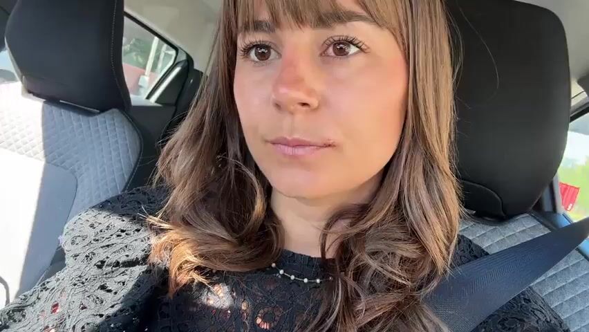 A woman with brown hair and bangs, wearing a black lace top and a seatbelt, is sitting in the driver's seat of a car. She looks at the camera and makes a kissing face, holding a strand of her hair. She is wearing gold earrings and a ring on her left ring finger. The car interior is visible, with patterned seats. The view out the window is blurred.