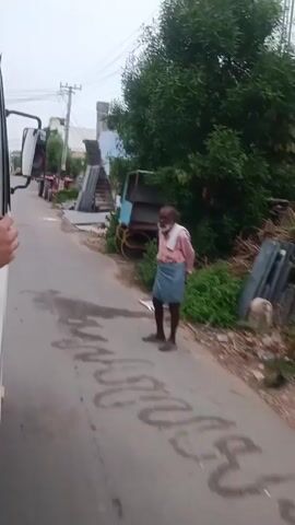 The video shows a street scene from the perspective of someone in a vehicle. A man wearing a pink shirt and a blue lungi stands on the right side of the road, facing away from the camera. He has a beard and is holding his hands out. To his left, there is a blue cart with a canopy and some yellow wheels, along with piles of wood and stone slabs. Several tractors are parked in a line on the left side of the road. In the background, there are houses and trees. The sky appears overcast. There are puddles of water on the road, suggesting it may have rained recently. The vehicle the camera is in is moving forward, and a hand is visible resting on the open window frame.