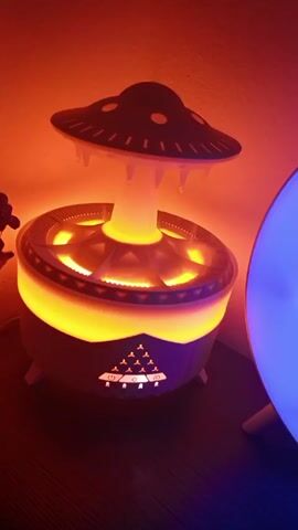 Here is a concise description of the video:

The video shows a humidifier with a unique design, shaped like a UFO. The humidifier is illuminated with a warm orange light, creating a cozy atmosphere. The room also features a blue light source in the background. The humidifier seems to be in operation, releasing mist from its top.  The overall setting appears to be indoors on a wooden surface.