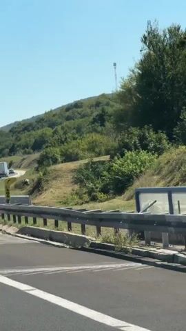The video is taken from a vehicle traveling on a highway. The road curves to the left, with a metal guardrail on the right side. Beyond the guardrail, there is a grassy slope dotted with trees. Two tall communication towers are visible on the crest of the hill, one taller than the other. The sky is clear and blue, indicating a sunny day. On the right side of the road, near the guardrail, a yellow SOS phone booth is visible. A truck is seen in the distance on the highway, moving away from the camera. The video is in landscape orientation and appears to be captured by a handheld device due to slight camera shake.
