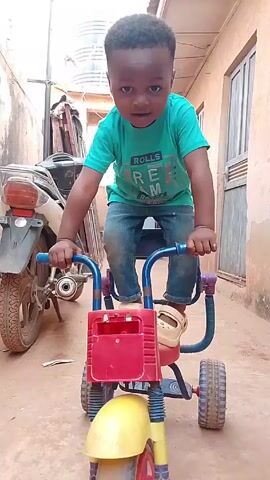A young child is standing astride a small, colorful tricycle in an outdoor setting. The child is Black, with short, dark hair, and is wearing a green t-shirt, blue jeans, and light-colored sandals. The tricycle has a red storage compartment in the front and a yellow mudguard over the front wheel. Behind the tricycle is a parked motorcycle and a building with a closed door and window. Another person, mostly out of focus, wearing a pink sweatshirt appears briefly in the foreground. The child seems to be posing or pretending to ride the tricycle. The ground appears to be made of dirt or packed earth.