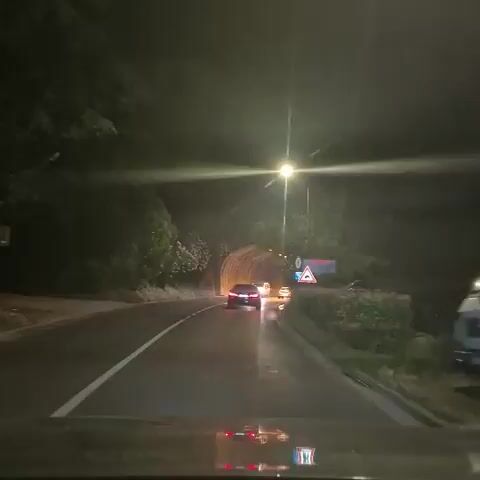 The video shows a nighttime view from the perspective of a vehicle driving towards a tunnel entrance. The road is wet, reflecting the headlights of the vehicles ahead and streetlights. Several cars are visible driving towards and into the tunnel. A motorcycle passes on the left side of the road. The tunnel is illuminated by a series of lights along its interior. Road signs are visible on the right side of the road, including a warning sign for a hump or dip in the road and a pedestrian crossing sign. Trees and foliage line the sides of the road leading up to the tunnel.