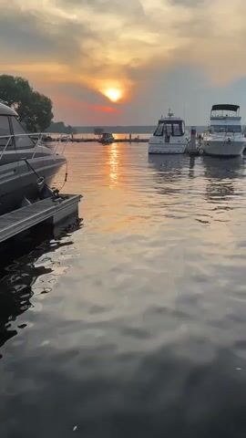 The video displays a marina at sunset. Several boats are docked, with one in the foreground on the left, partially obscuring the view. The sun is low on the horizon, casting a golden glow across the water and reflecting on the clouds. The sky is a mix of orange, pink, and gray hues. The water appears calm with gentle ripples. The environment suggests a peaceful, natural setting. There are no people visible in the video.
