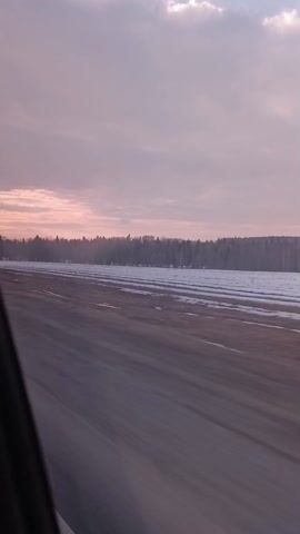 The video shows a car driving on an asphalt road during a dusky evening. The road is smooth and wet, reflecting the sky. On both sides of the road are snowy, frozen fields, with a dense forest lining the horizon. The sky is filled with pale pink and grey clouds, suggesting sunset or sunrise. The scene is filmed from inside the vehicle, with a small portion of the car's window frame visible on the left edge of the frame. The video lacks the sound of the car or any other obvious sounds. The camera is steady, indicating the car is moving smoothly along the road.
