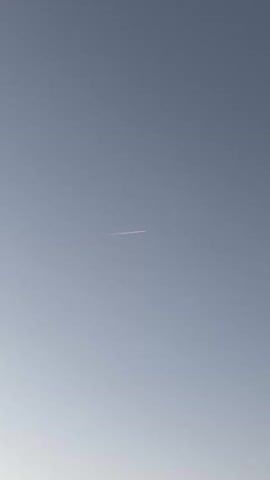A contrail, likely from an airplane, is visible in a clear sky. The contrail is thin and extends horizontally across the frame, with a slightly pinkish hue, suggesting it is illuminated by the sun. The sky is a pale blue, fading to a lighter shade towards the bottom of the frame. There are no other discernible objects or actions in the video.