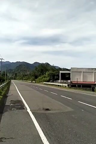 The video shows a gray car driving on a paved highway. The road has two lanes separated by a double yellow line, and a solid white line marks the edge of the road. On either side of the road are grass and trees. Mountains can be seen in the distance under a partly cloudy sky. A gray building is visible on the right side of the road. The camera appears to be stationary, capturing the car as it drives away. Power lines run along the left side of the road. There are some patches of what looks like dried water on the left side of the road.