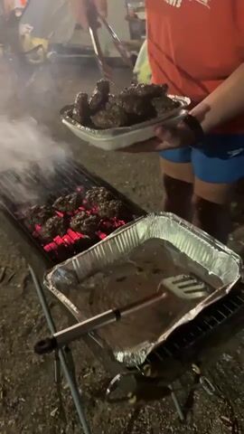 A person wearing an orange t-shirt and blue shorts is grilling burgers over hot coals. They are using tongs to flip the burgers on the grill. A foil tray is to the right of the grill, and another foil tray filled with cooked burgers is being held by the person. The scene appears to be outdoors at night, with some smoke rising from the grill.
