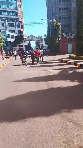 The video depicts an outdoor scene on a sunny day, showing a paved road with a group of people and a cyclist moving towards an entrance gate labeled "CHUKA UNIVERSITY."  Several individuals are walking along the road, some wearing red shirts and others in casual attire. A person on a bicycle is seen cycling in the same direction as the pedestrians. The shadows of the individuals and surrounding structures are visible on the road. In the background, there are tall buildings, including one under construction with a crane, and some greenery. The video appears to be shot from a ground-level perspective, capturing the movement and activity on the road leading to the university entrance.