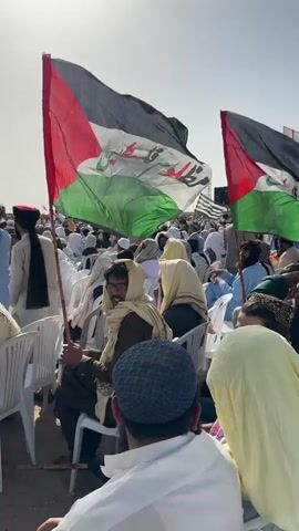 The video captures a large outdoor gathering of men, primarily adults, seated in white plastic chairs under a bright, clear sky in Pishin, Pakistan. The scene appears to be a public rally or event.

Two large Palestinian flags are prominently displayed in the foreground, held upright by wooden poles. These flags feature the black, white, green, and red horizontal stripes, with Arabic script "فلسطين" (Palestine) clearly visible in white on the green stripe. Another flag with similar design is visible further back on the right.

The crowd extends into the background, filling the open, sunlit space. Most attendees are dressed in traditional attire, including turbans, kufis, and shawls, consistent with the region. Many men have beards. Their backs are mostly towards the camera, but some individuals in the foreground are visible from the side or front.

The event takes place on what appears to be a dusty or arid ground surface. The bright sunlight indicates it is midday, with minimal shadows. In the far background, a large speaker or structure is partially visible. The attendees appear attentive, suggesting they are listening to a speaker or participating in a planned program. One individual in the foreground is seen holding a smartphone. The overall atmosphere suggests a focused and organized demonstration of solidarity.