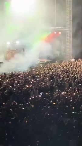 A wide shot captures a lively concert with a large crowd of people in the foreground. Many audience members are holding up their phones, illuminated by the stage lights and creating a sea of small lights. The stage in the background is filled with smoke and haze, illuminated by bright green and red stage lights. A lone figure, presumably a performer, stands on the stage, silhouetted against the lights. The overall atmosphere appears energetic and exciting.
