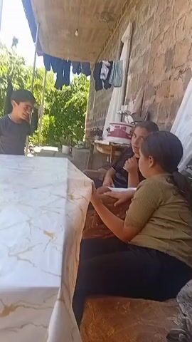 Two young girls and a young boy are present. The girls are seated on a couch or bench. One girl, in the foreground, is wearing a light green short-sleeved shirt and black pants, and she is looking down at a phone in her hands. The second girl, behind her, is wearing a dark t-shirt and is also looking at a phone. The boy, standing, is wearing a dark blue t-shirt and jeans, and he is looking towards the camera. The scene appears to be outdoors, possibly a courtyard or patio, with a stone wall in the background, clothes hanging on a line overhead, and trees visible. A table covered with a white and gold marble-patterned tablecloth is in the foreground. The boy is positioned behind the table, facing the girls.
