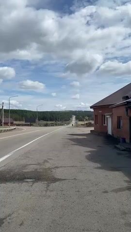 The video shows a straight, two-lane asphalt road extending into the distance, disappearing over a slight hill. The sky is mostly blue with puffy white clouds. A line of trees runs along the horizon. On the right side of the road is a low, brick building with a brown roof. A few parked cars are visible on the left side of the road, and a person is walking on the road in the distance towards the camera. There are streetlights along the road. The shoulder of the road near the camera is cracked and has some puddles.