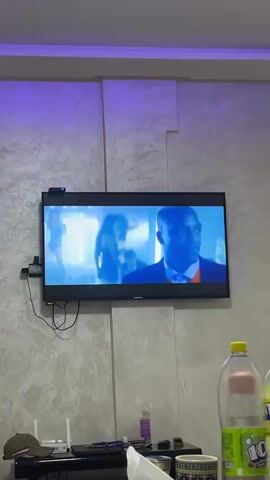 The video shows a television screen displaying a music video. The scene transitions between close-ups of a Black man in a suit and a White man with two Black women, and a group of people dancing. The background of the TV screen is blurred and appears to be a club or party setting. The room where the TV is located has textured walls with a purple light illuminating the top portion. In the foreground, on a table, are a plastic bottle of soda, a white tissue, a cup, and other miscellaneous items.