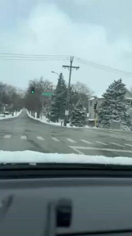 The video depicts a view from inside a car stopped at an intersection on a snowy day. The street ahead is covered with slush and patches of snow. Trees and houses along the street are also dusted with snow.  A traffic light ahead is green, while a traffic light perpendicular to the view is red. There are crosswalk markings on the road, partly obscured by the snow. Power lines run above the street.  A street sign reading "Maplewood Rd" is visible near the traffic light. The dashboard of the car is visible at the bottom of the frame.