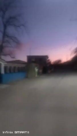 The video shows a blurry, slightly elevated view of a street scene during twilight. The sky is a mix of purple and orange hues. On the left side of the frame, there's a building with a blue wall. Further down the street, there's a small store with its lights on, and several people are gathered outside with bicycles. Across the street is another building. Power lines are visible overhead. The ground is dusty and gray. The quality of the video is low, resulting in a lack of detail. The bottom of the frame displays the text "Infinix SMART 8."