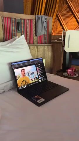 A dark-colored laptop rests on a white bed with a white pillow. The laptop screen displays a video, featuring a blond man in a yellow sweatshirt with "LOS ANGELES" text, speaking in front of a house. To the right of the video on the screen is a vertical sidebar with text and thumbnails. Behind the bed is a wooden headboard with a rectangular panel featuring a red, pink, and white striped textile pattern. To the right, a dark gray garment hangs on a hook on a wooden wall. Further right, a light-colored surface holds a white plate with sliced pink fruit (possibly dragon fruit) and a small, dark vase with a pink flower. The background wall has a textured, possibly woven, pattern. A buzzing sound is audible.
