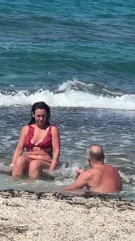 Two people, a woman and a man, are sitting in shallow water near a beach. The woman is wearing a red bikini. The man is facing away from the camera, and his back appears to be sunburned. The water is clear and blue, with waves breaking on the shore. The beach is covered in sand and some seaweed. The woman appears to be looking at the man, and he is gesturing with his hands.