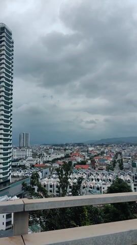 The video presents a panoramic view of a coastal city and bay from a high vantage point, likely a balcony or rooftop. The scene is set under a heavily overcast, cloudy sky, giving the landscape a somewhat muted appearance.

In the initial frames, a modern, tall skyscraper with a distinctive white and dark architectural pattern dominates the left side of the view. Below and stretching into the distance, numerous residential and commercial buildings populate the city, some featuring uniform, multi-story designs with angular roofs.

As the camera pans to the right, the tall building comes into clearer view, revealing the text "DIAMOND" prominently displayed near its top. The expansive body of water, a bay, becomes more visible, dotted with a significant number of boats of varying sizes, including what appear to be cargo ships and smaller vessels. Mountains form the distant horizon, fading into the cloudy sky.

The pan continues further right, showcasing more of the bustling bay with its maritime activity, and the distant mountainous coastline under the persistent gray sky. A metal railing is visible in the immediate foreground throughout the video, indicating the elevated position of the camera. The overall impression is that of a busy coastal area, possibly a port city, on a gloomy, overcast day. The setting aligns with Hong Gai Ward, Vietnam, which is part of Hạ Long City and overlooks Hạ Long Bay.