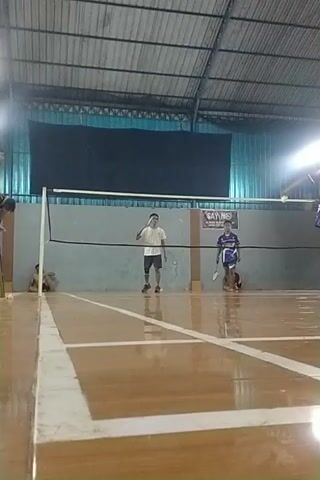 The video shows an indoor badminton court. There are at least four people present. Two individuals appear to be playing badminton, one in a blue and white shirt and shorts, and another in a black t-shirt and shorts. A third person in a white t-shirt and shorts is standing on the court, and a fourth person is partially visible in the foreground. The court has wooden flooring with white boundary lines. A net divides the court. The lighting appears to be from overhead fixtures, creating some glare. The walls of the indoor facility are visible, with some netting and metal structures in the background. The camera angle is angled and the video is partially rotated.