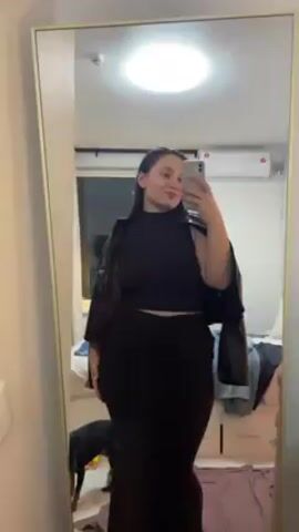 A young woman is taking a selfie in a full-length mirror. She is wearing a dark brown, two-piece outfit consisting of a sleeveless crop top and a long, fitted skirt. A dark brown leather jacket is draped over her shoulders. She poses slightly, looking at the camera. A dog is briefly visible near the bottom left corner of the frame. The room appears to be a bedroom with a bed, a nightstand, and a window visible in the background. There's a small air conditioning unit near the ceiling.