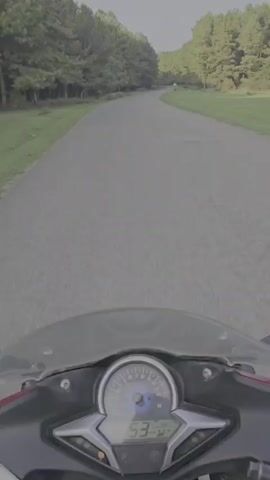 A first-person perspective video shows a motorcycle being ridden on a paved road through a wooded area. The motorcycle's speedometer is visible, showing a speed of 54 km/h. The road is a two-lane asphalt road with a grassy shoulder on either side. Tall trees line the road, and the sky is overcast. The motorcycle appears to be red and black. The rider is wearing a helmet. The video shows the motorcycle moving forward at a consistent speed. There are no other vehicles or people visible on the road.