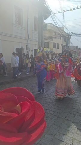 A short video depicts a parade on a cobblestone street in a town.  The parade participants include individuals in elaborate, colorful costumes with masks.  Some of the costumes incorporate fringe and intricate patterns.  Behind the masked figures, a group of children dressed in traditional clothing with hats and shawls follow along.  The buildings lining the street are primarily white with some featuring commercial signage.  A few bystanders watch the parade from the sidewalks.  Overhead power lines are visible. The lighting is bright, creating glare in the sky, suggesting a sunny day.  A corner of a red decorative object appears in the foreground.