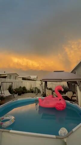 This video shows a backyard scene at dusk. The sky is filled with dramatic, orange and gray clouds. In the foreground, a round, above-ground swimming pool is filled with blue water. A large, inflatable pink flamingo float is in the pool, and a smaller, circular pool float is near the edge. A white fence encloses the backyard, and behind it, several small buildings or mobile homes are visible. A gazebo with outdoor furniture is also present. The overall impression is one of a serene, possibly end-of-day, atmosphere. There are no people visible in the video.