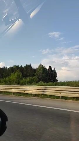 The video shows a car driving on a highway on a sunny day. A guardrail runs along the side of the road, separating it from a dense forest with a mix of deciduous and coniferous trees. The sky is blue with scattered white clouds. The camera pans slightly upwards, focusing more on the sky and treetops. The video appears to have been taken from inside the moving car. Music is audible in the background.