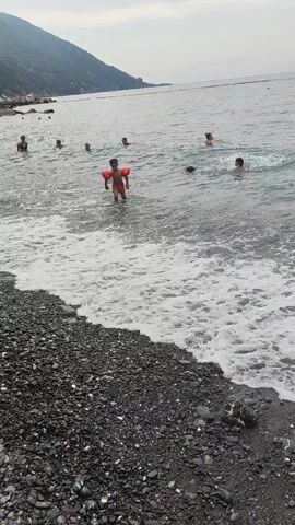 The video shows a rocky beach with a body of water. Several people are in the water, including a child wearing orange arm floats. The child is standing in shallow water near the shore, facing away from the camera. The sky is overcast and a distant hill is visible in the background. The waves are small and gentle. The overall scene depicts a calm day at the beach with people enjoying recreational activities in the water.