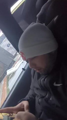 A man in a car is looking down at something in his hands, possibly a phone or a small object. He is wearing a gray beanie with a red logo and a black Nike jacket. He has a beard and appears to be in his late 20s or early 30s. The car window shows a blurry outdoor scene with snow or slush present. The interior of the car is dark. There is music playing.