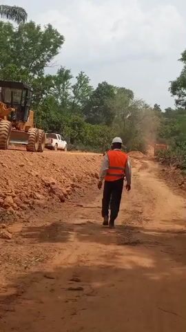Three men walk down a dirt road. The man in front is wearing a white helmet and an orange reflective vest. The other two men are wearing casual clothes. There's a grader parked on a dirt berm to the left of the road. A white pickup truck is parked further down the road on the left. There are trees and vegetation on both sides of the road, and the sky is cloudy.