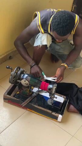 A dark-skinned man is crouched down on a beige tiled floor, using scissors to cut a piece of black fabric on an old-fashioned sewing machine. The man is wearing a black and yellow basketball jersey and frayed denim shorts. He has a black wristband on his left wrist and a gold bracelet on his right. The sewing machine is housed in a wooden base painted with colorful stripes. The machine itself has been painted in a variety of colors. A pair of pliers is sitting on the base of the machine. To the right of the machine, there is a brown wallet on the floor. The man is focused on his task, carefully cutting the fabric. A closed wooden door is visible in the background. The scene is well-lit.