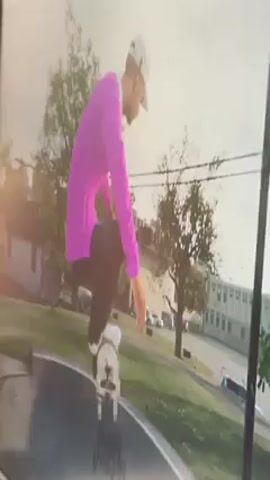 A person wearing a pink long-sleeved shirt, dark pants, white shoes, and a white hat is skateboarding in a skate park. The person starts at the bottom of a curved ramp and skates up, balancing at the top, before rolling back down. The skate park is outdoors with grass and trees in the background, as well as buildings and parked cars. The video appears to be a recording of a screen displaying a video game.
